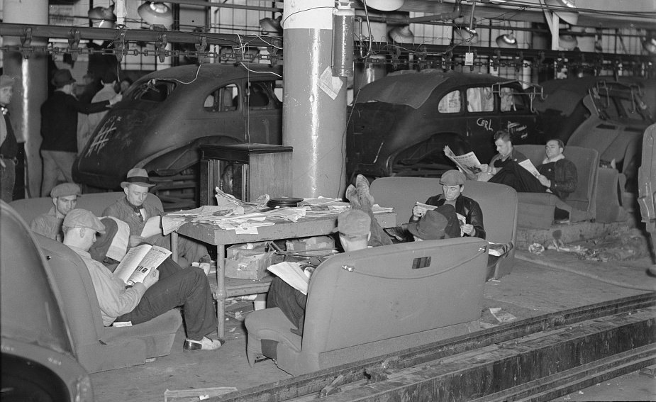 Photo showing workers sitting down at a strike with half-built automobiles behind them.